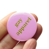 Pale pink round pinback button that reads GAY APPAREL in moss green serif text.  Button is held in a hand.