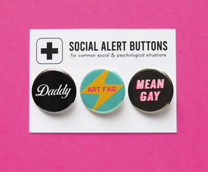 Three pinback buttons lined up on a SOCIAL ALERT BUTTONS backing card. Reading Left to Right: A black pinback button that reads Daddy in a white script font, A teal button with a yellow lightening bolt that reads ART FAG in pink, and a black button with tiny white dots that reads MEAN GAY in light pink with a dark pink drop shadow