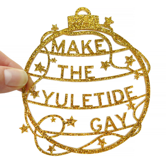 Round gold glitter acrylic laser cut holiday ornament that reads MAKE THE YULETIDE GAY within a classic ornament shape, with stars and garlands around it.