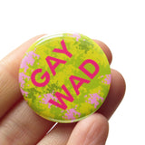 Round yellow green patterned pinback button that reads GAY WAD in dark pink text. Badge is held in a hand.