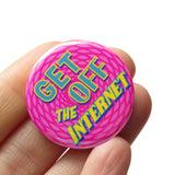 A round pink pinback button with a circular background pattern t that reads GET OFF THE INTERNET in yellow & teal text. badge is held in a hand.