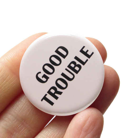 A round off-white pinback button that reads GOOD TROUBLE in black text. Button is held in a hand.