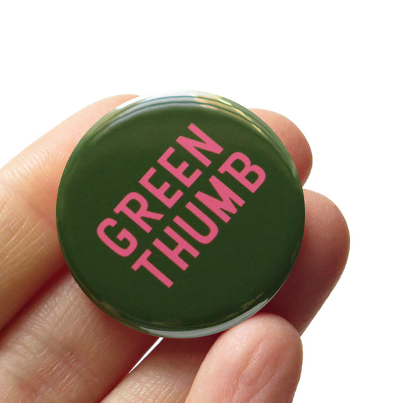 A round mossy green button with rosy peach text that reads GREEN THUMB. Button is held in a hand