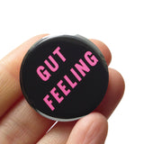 Round black pinback button with pink text reading GUT FEELING on two lines. Badge is held in a hand.