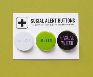 Three round pinback buttons lined up on a SOCIAL ALERT BUTTONS backing card. Reading left to right: GHOST in white & gray, GOBLIN in greens & CASUAL WITCH in black & lavender.  