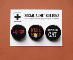 Three round pinback buttons lined up on a SOCIAL ALERT BUTTONS backing card. Reading left to right: VAMPIRE in black & red, WERE WOLF in brown & black, and SCARDEY CAT in black & white.