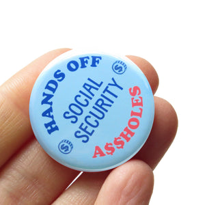 A round light blue pinback button with light dark blue text reads HANDS OFF SOCIAL SECURITY with two little illustrated coins with dollar signs on them, and A$$HOLES in neon red at the bottom. Button is held in a hand.