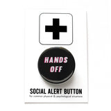Round black pinback button reading HANDS OFF in white text with pink drop shadow. Badge is on a Social Alert Button backing card.