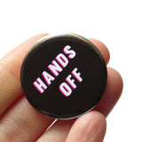 A round black pinback button that reads HANDS off in white text with a pink drop shadow. Badge is held in a hand.