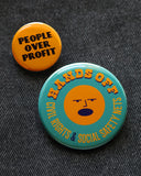 A big button and a small button pinned to black denim. The big round teal pinback button reads HANDS OFF Civil Rights & Social Safety Nets yellow-orange text,  with an angry shouting face in the center of the button. The smaller yellow-orange button reads PEOPLE OVER PROFIT in black text.