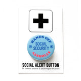 A round light blue pinback button with light dark blue text reads HANDS OFF SOCIAL SECURITY with two little illustrated coins with dollar signs on them, and A$$HOLES in neon red at the bottom. Button is on a social alert button backing card.