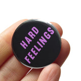 Round black 1.25" pinback button that reads  HARD FEELINGS in lavender text. Button is held in a hand.