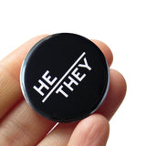 Round pinback button with black background which reads HE/THEY  on an angle in white text, in a san serif font. Button is held in a hand.