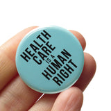 Round pinback button that says HEALTHCARE IS A HUMAN RIGHT. Black text on a hospital blue-green background. Button is held in a hand.
