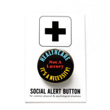 a round black pinback button with yellow, red and cyan text that reads HEALTHCARE IT'S A NECCESSITY, NOT A LUXURY! Button is pinned to a Social Alert Button backing card.