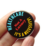 a round black pinback button with yellow, red and cyan text that reads HEALTHCARE IT'S A NECCESSITY, NOT A LUXURY! Button is held in a hand.