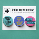 3 round multi-colored pinback buttons lined up on a SOCIAL ALERT BUTTONS backing card. Reading left to right: Healthcare Is a Human Right, Public Health Over Corporate Wealth, Save Medicaid
