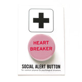 Round light pink pinback button that reads HEARTBREAKER in dark pink on two lines. Badge is on a Social alert button backing card.