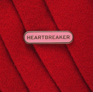 Capsule shaped enamel pin that says HEARTBREAKER. Silver text and outline on a pink enamel background. On red felt jacket.