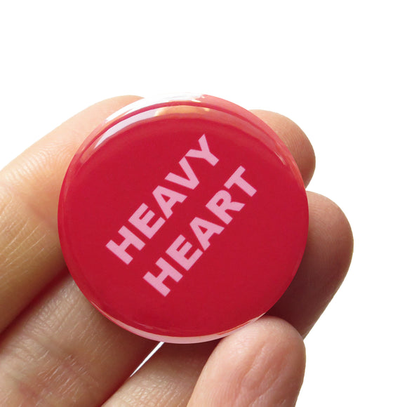 Round dark pink magenta pinback button that reads HEAVY HEART on two lines in a chunky light pink font. Badge is held in a hand.