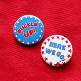 Two round pinback buttons, one is blue with white stars around the edge, reading Buckle Up! in red text. The other Is white with red stars around the border and reads HERE WE GO! in blue text that starts to collapse visually. Both buttons are on red denim.