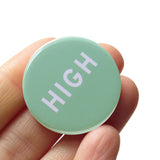 Round pinback button that says HIGH. White text on a mint green background. Badge is held in a hand.