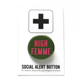 Round pinback button that HIGH FEMME. Pink text with a red shadow on an olive green background. Button is pinned to a Social Alert Button backing card