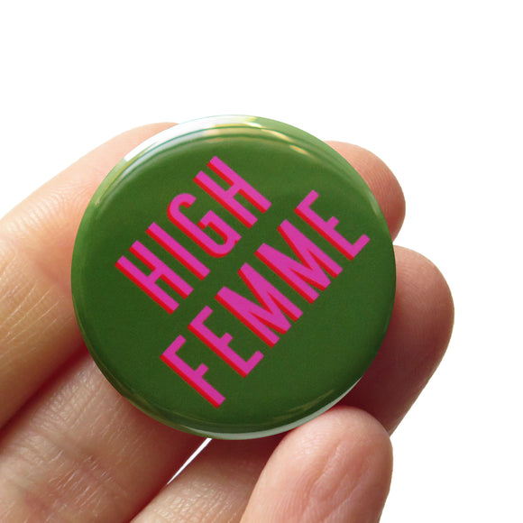 Round pinback button that HIGH FEMME. Pink text with a red shadow on an olive green background. Button is held in a hand.