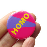Round pinback button with neon red-pin background with violet stripe which reads HOMO in bold yellow text. Button is held in a hand.