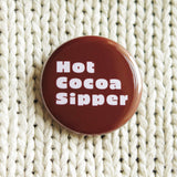 round chocolate brown colored 1..25" holiday themed pinback button that reads HOT COCOA SIPPER in a chunky white font. Button is pinned tp  white sweater