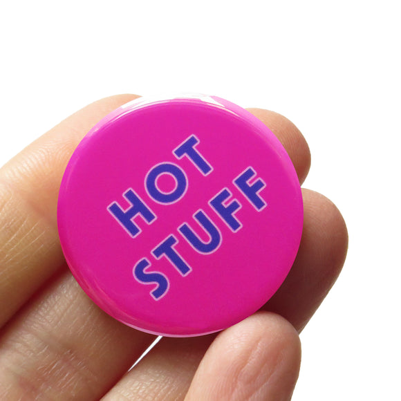  round bright pink pinback button that reads HOT STUFF in two lines in purple text. Button is held in a hand.