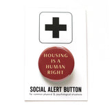 Round red-brown pinback button that reads HOUSING IS A HUMAN RIGHT in a cream color font. Pinback button is on a SOCIAL ALERT BUTTON backing card.