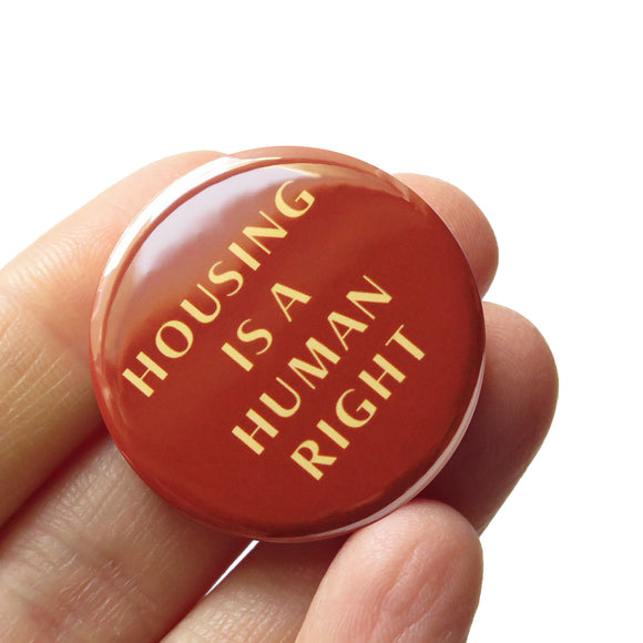 Round red-brown pinback button that reads HOUSING IS A HUMAN RIGHT in a cream color font. Hand in a white hand.