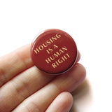 Round red-brown pinback button that reads HOUSING IS A HUMAN RIGHT in a cream color font. Hand in a white hand.