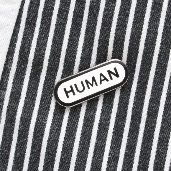 Capsule shaped hard enamel lapel pin with dark gray metal spelling HUMAN in a san serif font, with a dark metal outline. The pin is a diagonal strip jacket.