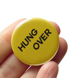 A round yellow-green pinback button that reads HUNG OVER in two lines in dark text. Badge is held in a hand
