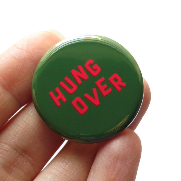 A round dark green pinback button that reads HUNG OVER in slightly askew neon red letters for a Christmas look.  Held in a hand, up close.