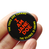 Round black pinback button that reads I AM JANE DOE  in neon orange text, and yellow curving text  reading: the tides are Turing, the silence is ending. The button is held in a hand.