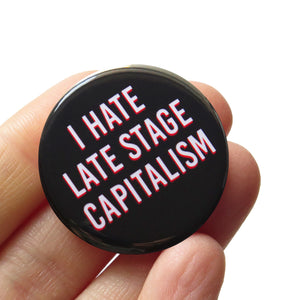 Round black pinback button that reads I HATE LATE STAGE CAPITALISM in white text with a red drop shadow. Pin is held in hand.