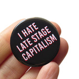 Round black pinback button that reads I HATE LATE STAGE CAPITALISM in white text with a red drop shadow. Pin is held in hand.