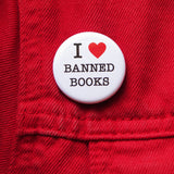 A round white pinback button that reads I LOVE BANNED BOOKS. Love represented by a red heart. Button is on a red denim jacket.