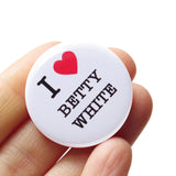 A shiny, round white pinback button with black serif text and a read heart, the button reads: I LOVE BETTY WHITE, with love being illustrated by a red heart. The button is held in a hand.