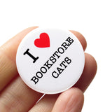  round white 1.25" pinback button that reads I LOVE BOOKSTORE CATS, love being represented by a red heart. Badge is held in a hand.