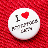  round white 1.25" pinback button that reads I LOVE BOOKSTORE CATS, love being represented by a red heart. Badge is pinned to a sweater