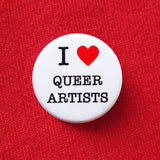 A round white pinback button that reads I LOVE QUEER ARTISTS in a black serif font, the love being indicated by a red heart. The button is pinned to a red sweatshirt