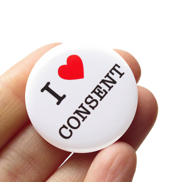A round white button that reads I LOVE CONSENT, love being a red heart. Buton is held in a hand.