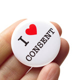 A round white button that reads I LOVE CONSENT, love being a red heart. Buton is held in a hand.