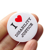 A round white 1.25" pinback button that reads I LOVE DISABILITY JUSTICE, with love being represented by a red heart. Badge held in a hand.