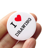 A round white pinback button that reads I LOVE DRAWING, love being represented by a red heart. Button is held in a hand.