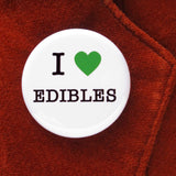 Round white button that reads I LOVE EDIBLES. Love is indicated with a green heart symbol, the remaining text is black. Button is pinned to rust velvet blazer lapel.